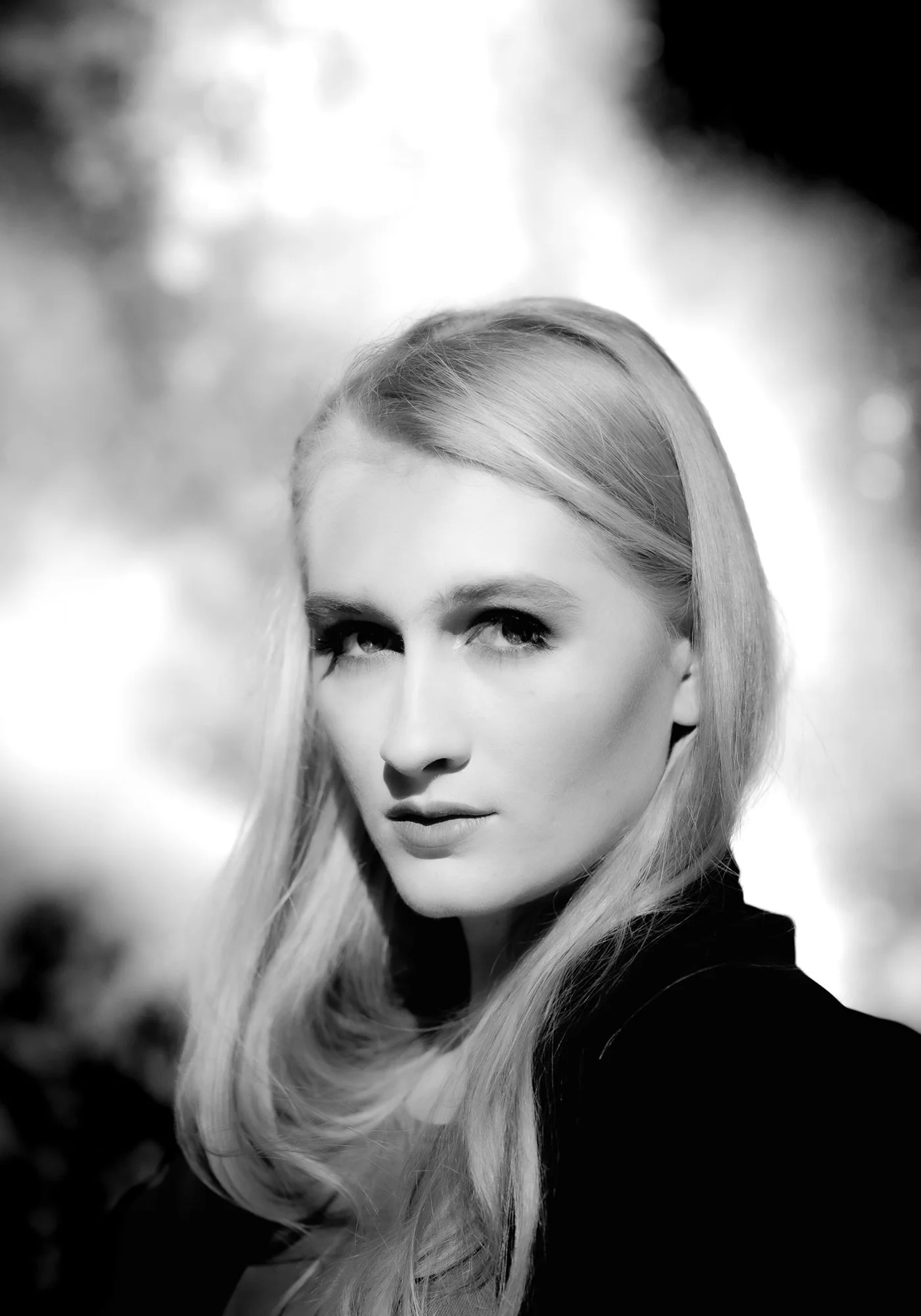 Black and white portrait of a blonde woman with long hair, gazing intently at the camera, outdoors with blurred trees in the background.