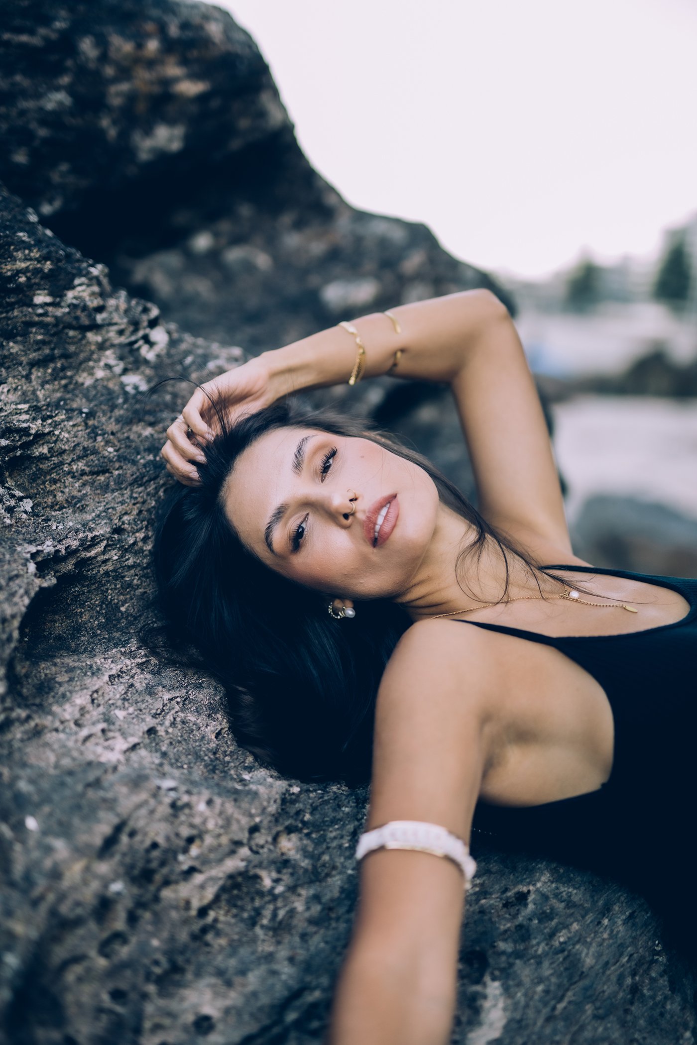 A young woman with long dark hair lying on a rocky outdoor surface, wearing a black tank top and jewelry, with her eyes partially closed and one hand resting on her head.