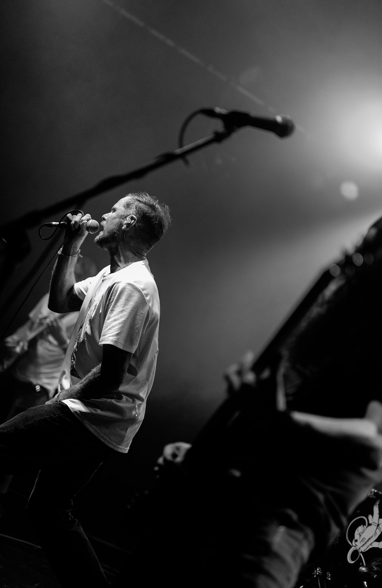 A black and white photo of a man singing into a microphone on stage, side view, with microphone stand in the foreground and an electric guitar partially visible.
