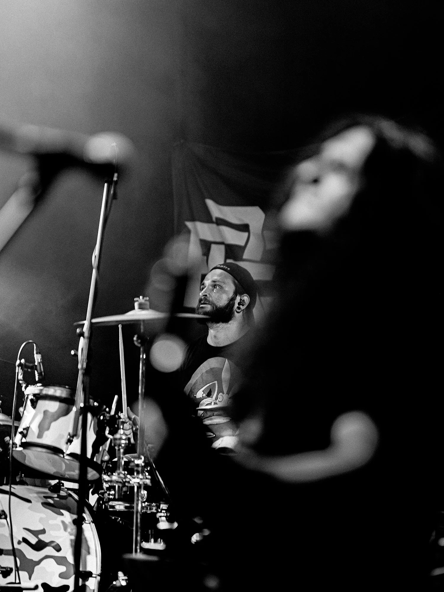 A drummer with a beard wearing a cap performing on stage with a camouflage drum set, black backdrop, and a partially visible band member in the foreground.