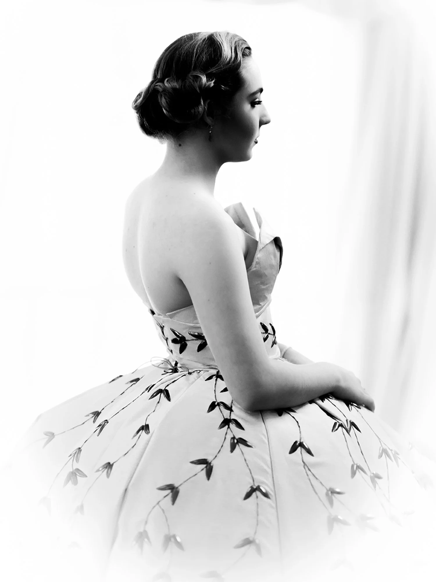 A young woman in a ball gown stands by a window as light comes through