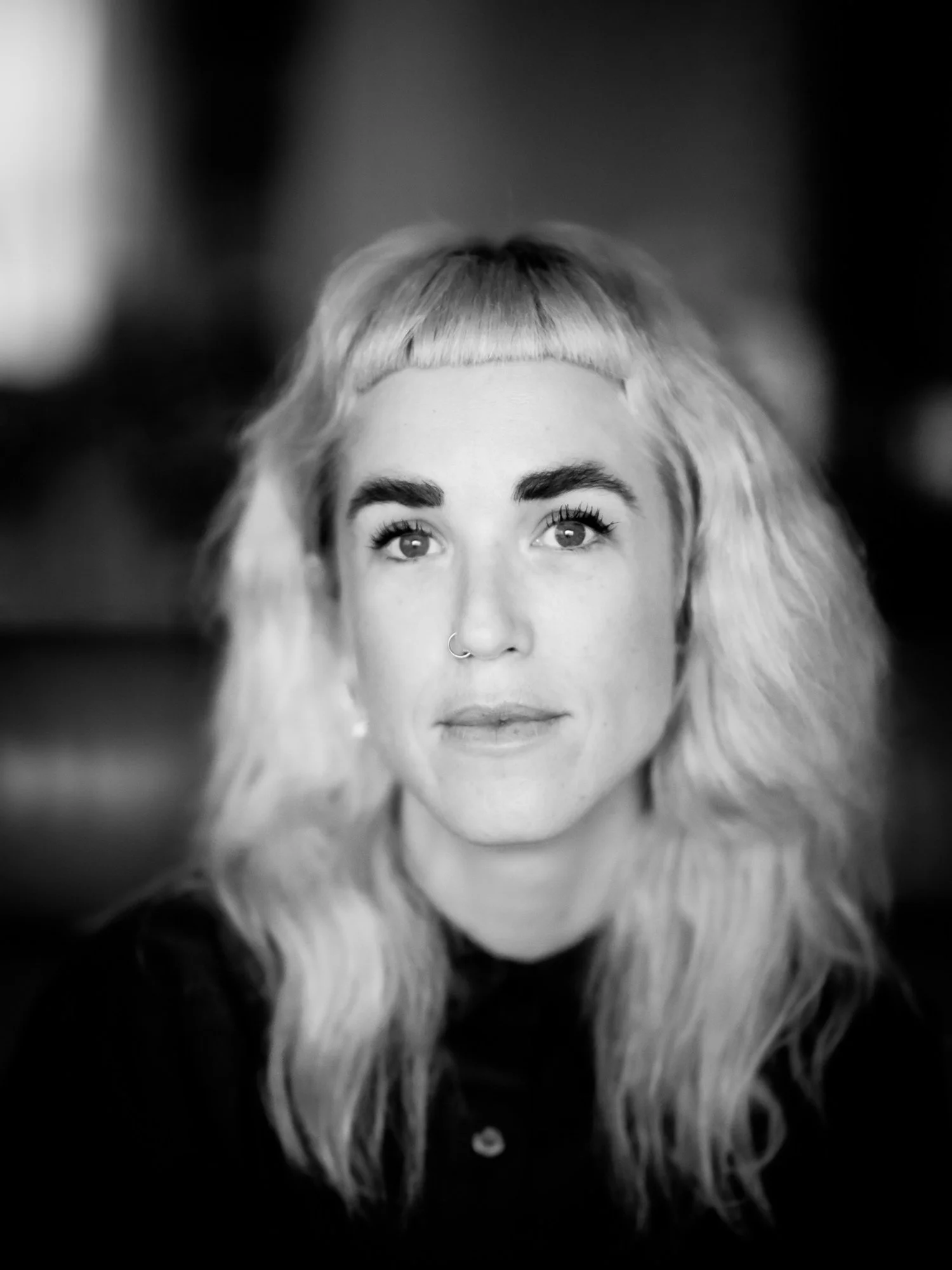 A black and white portrait of a woman with wavy blonde hair, prominent eyebrows, a septum piercing, and wearing a dark top, looking directly at the camera.