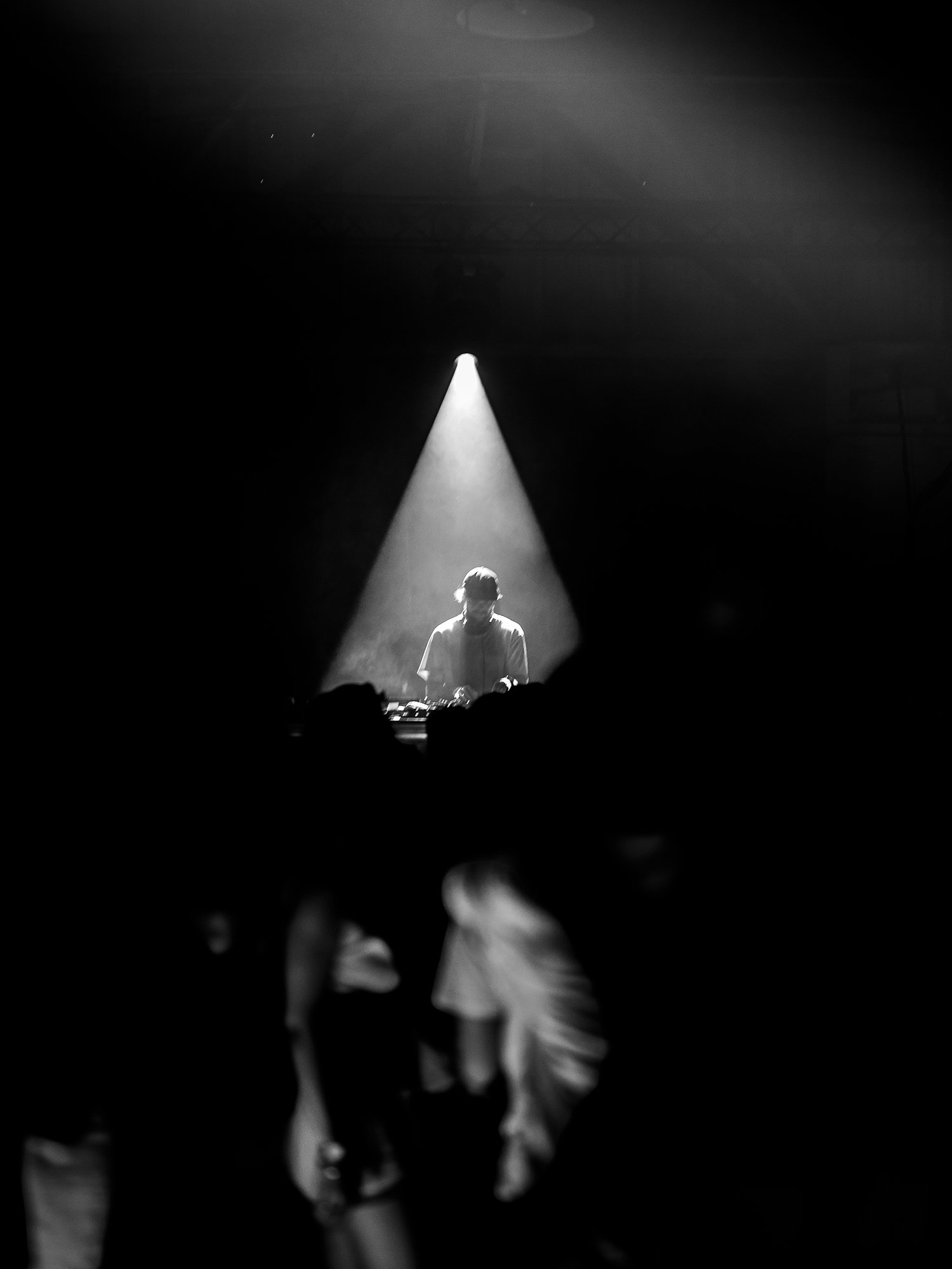 A DJ performing on stage under a spotlight at a concert with a dark background and audience silhouette in foreground.
