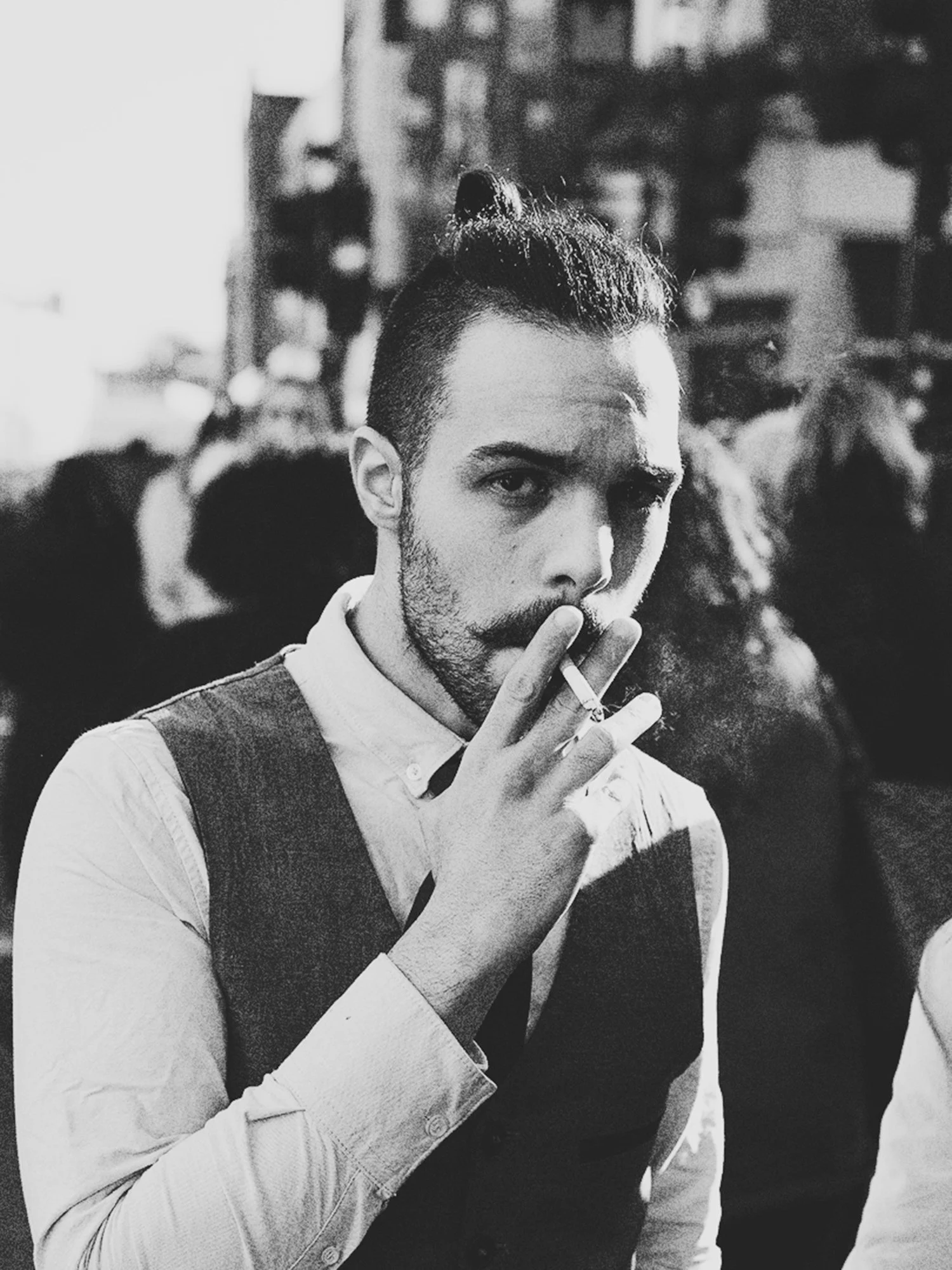 A black and white photo of a man with a beard and mustache, smoking a cigarette, wearing a button-up shirt and a vest, with a serious expression, in a crowded indoor setting.