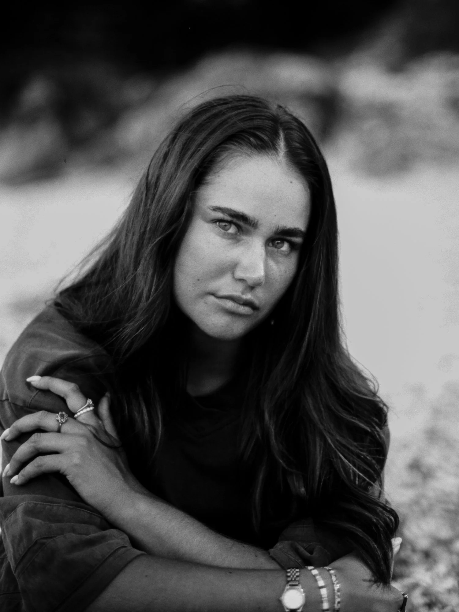 Black and white portrait of a young woman with long, dark hair, sitting outdoors with mountain scenery in the background, looking directly at the camera.