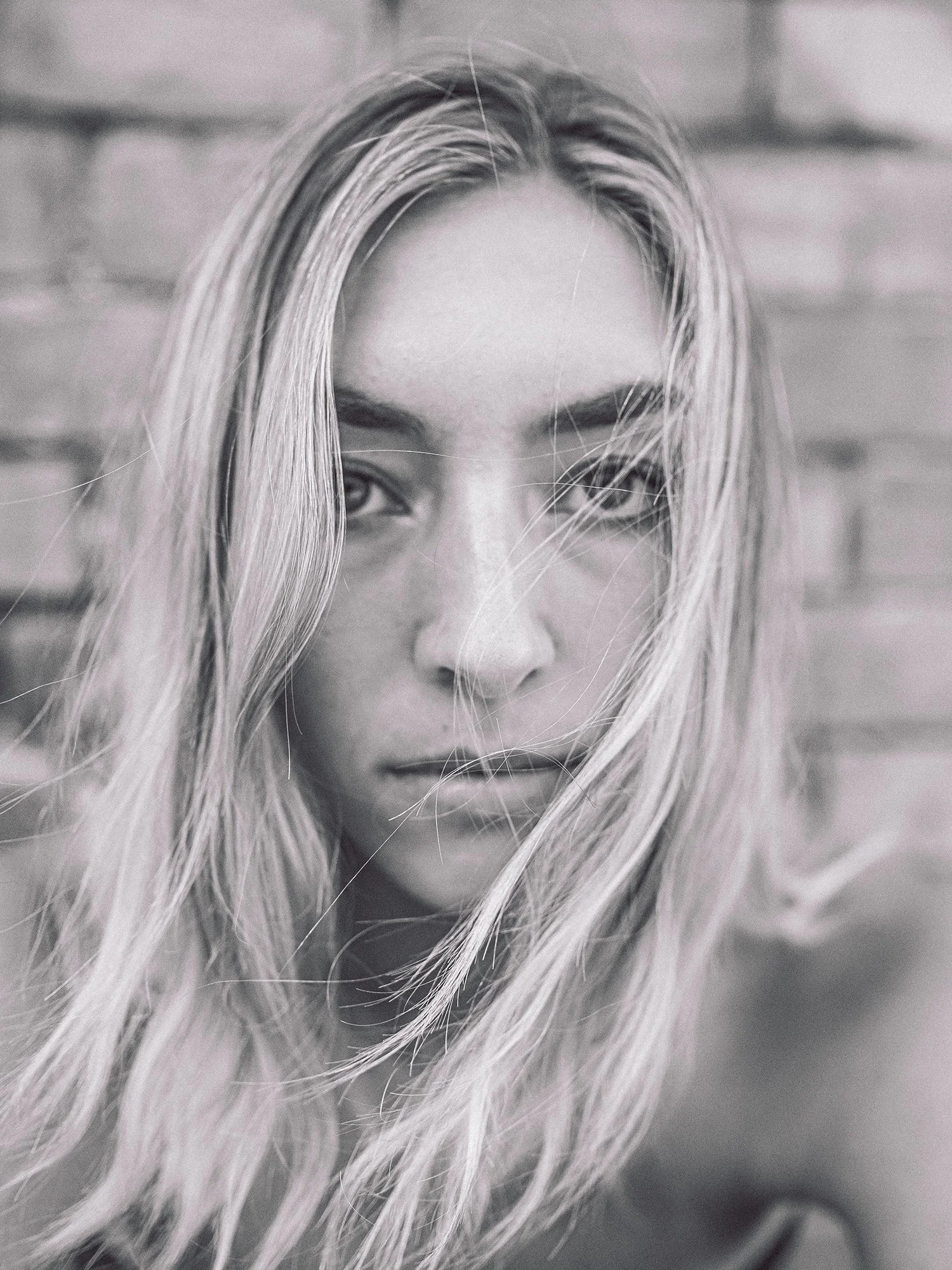 Close-up black and white photo of a young woman with long, wavy hair partially covering her face, looking directly at the camera with an intense expression.