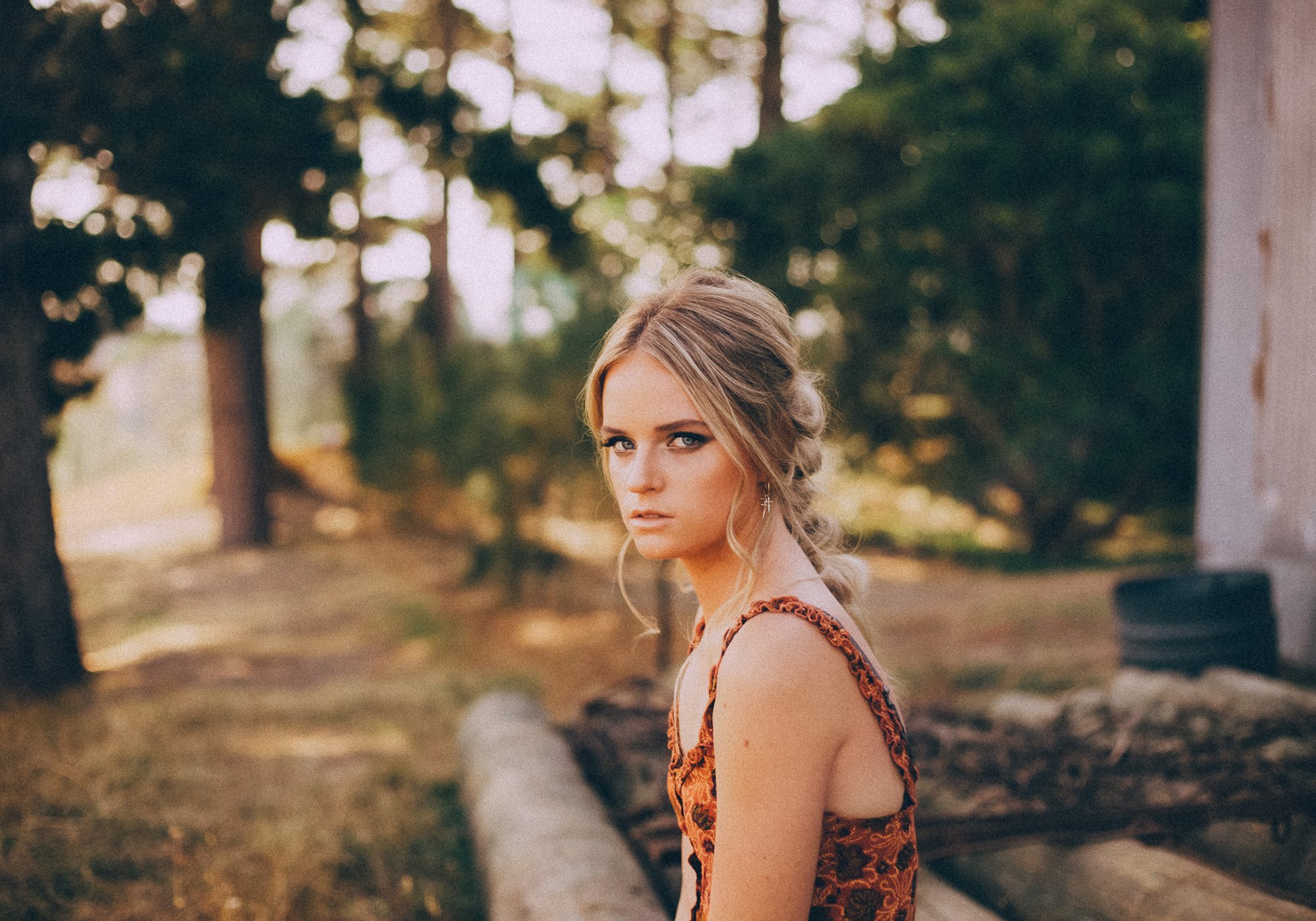 A young woman with blonde hair styled in a braid, wearing a sleeveless floral dress, stands outdoors with trees and a rustic wooden structure in the background, looking at the camera with a serious expression.