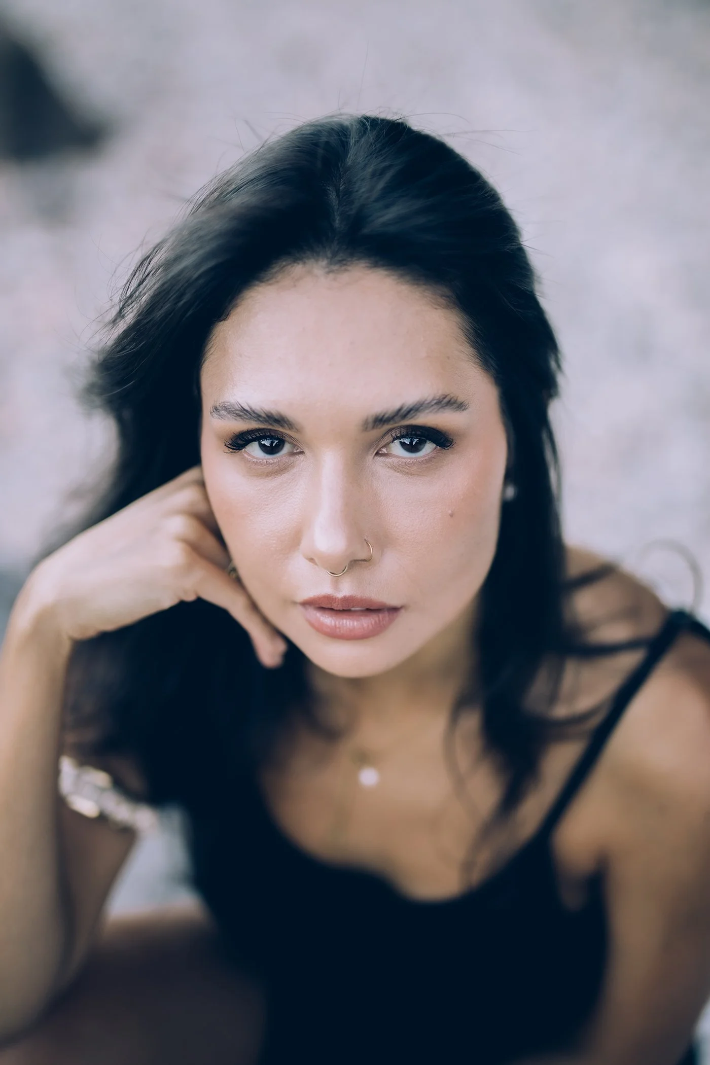 A woman with dark hair, wearing a black top, looking directly at the camera with a neutral expression, resting her hand near her face.