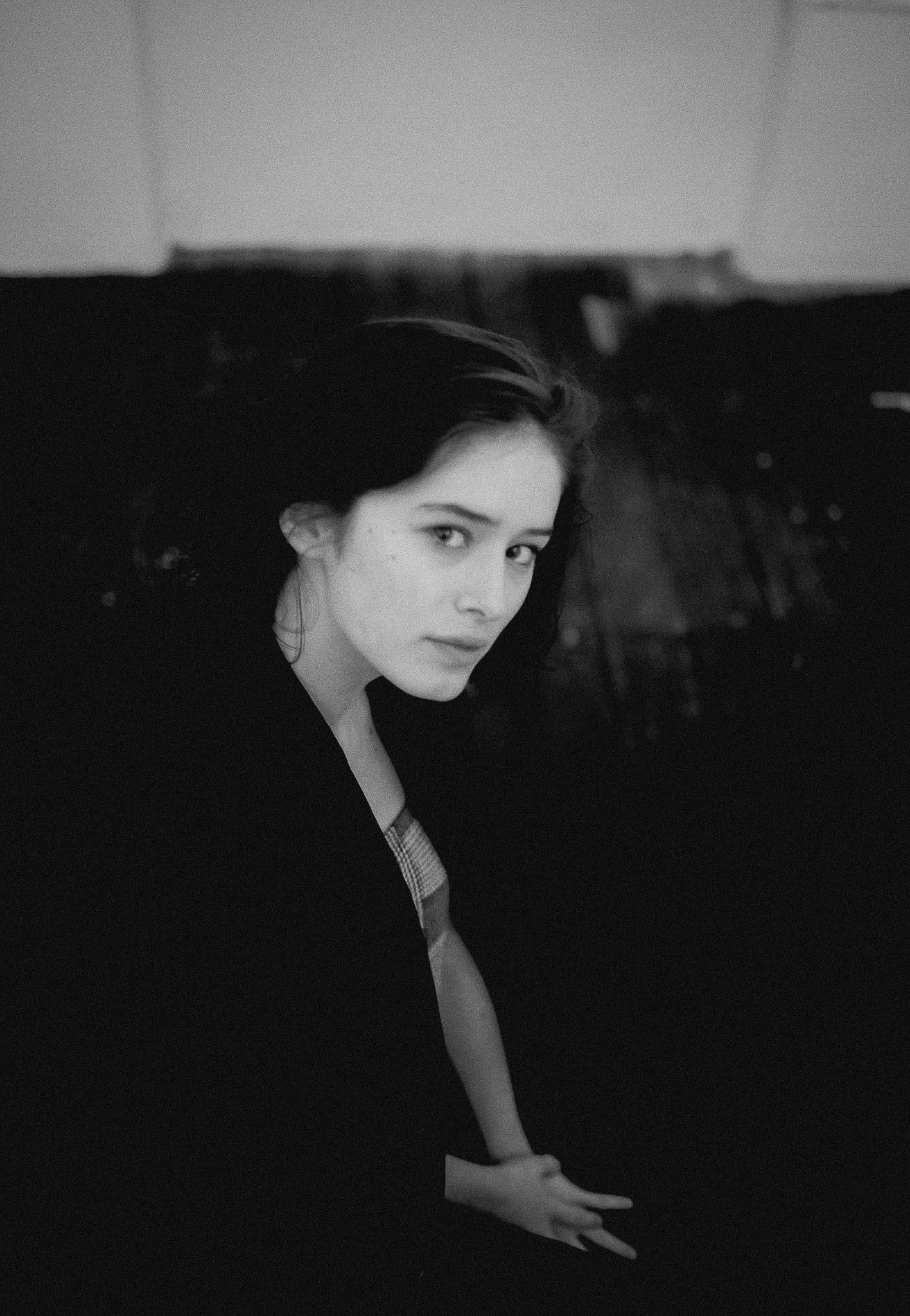 Black and white photo of a young woman with dark hair, looking over her shoulder at the camera, sitting against a dark background.