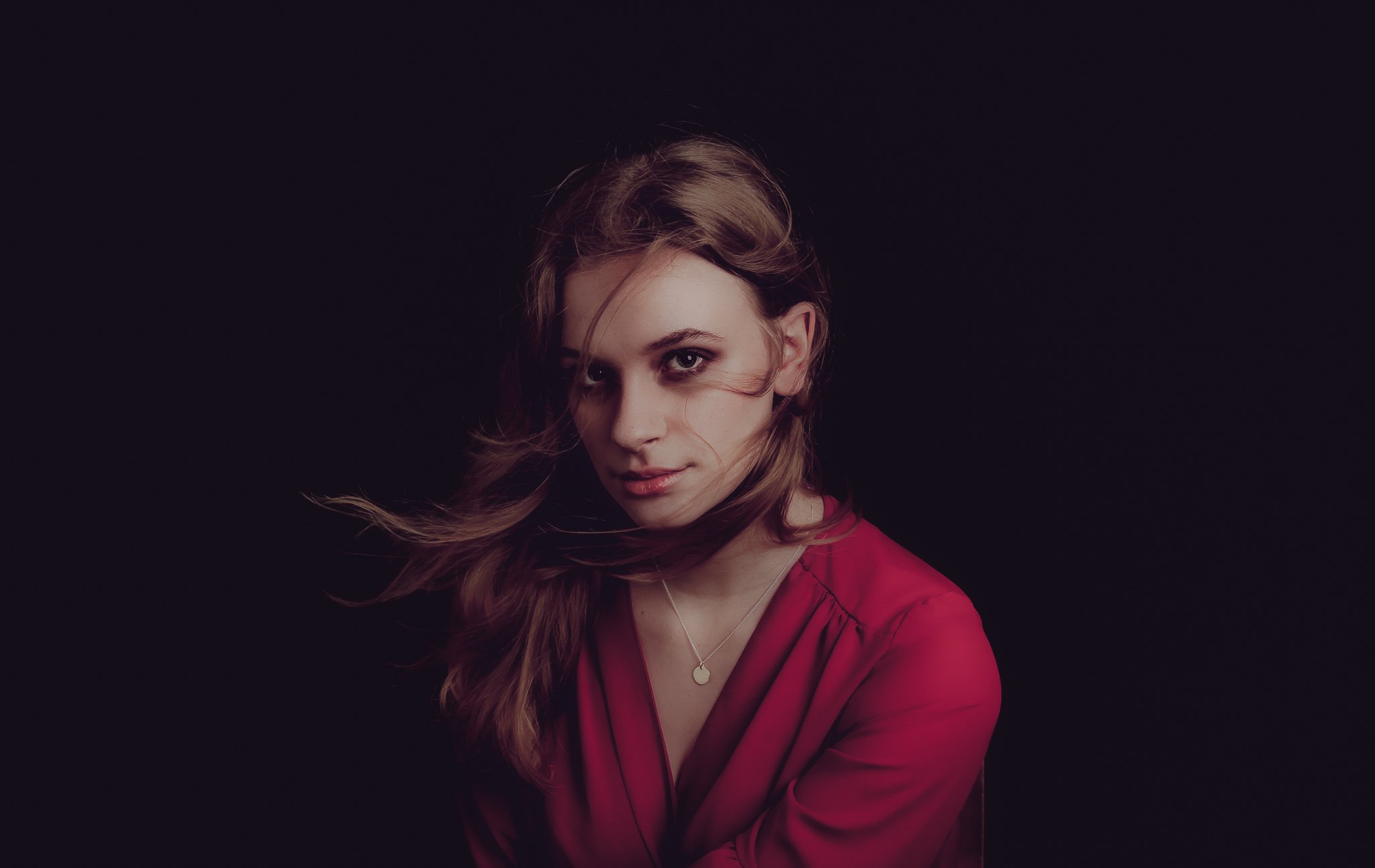 A woman with long hair wearing a red top, looking at the camera against a dark background.