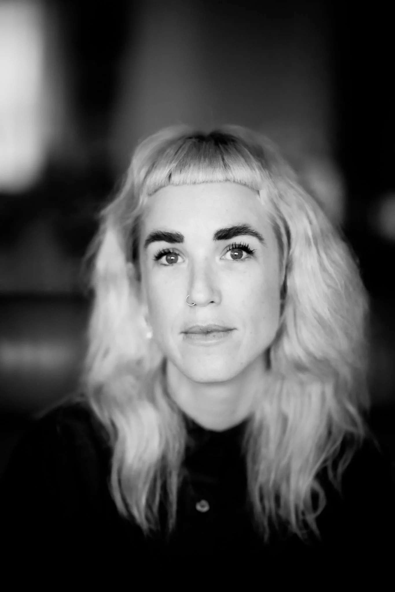 Black and white portrait of a woman with long, wavy blonde hair, a nose ring, and a calm expression.