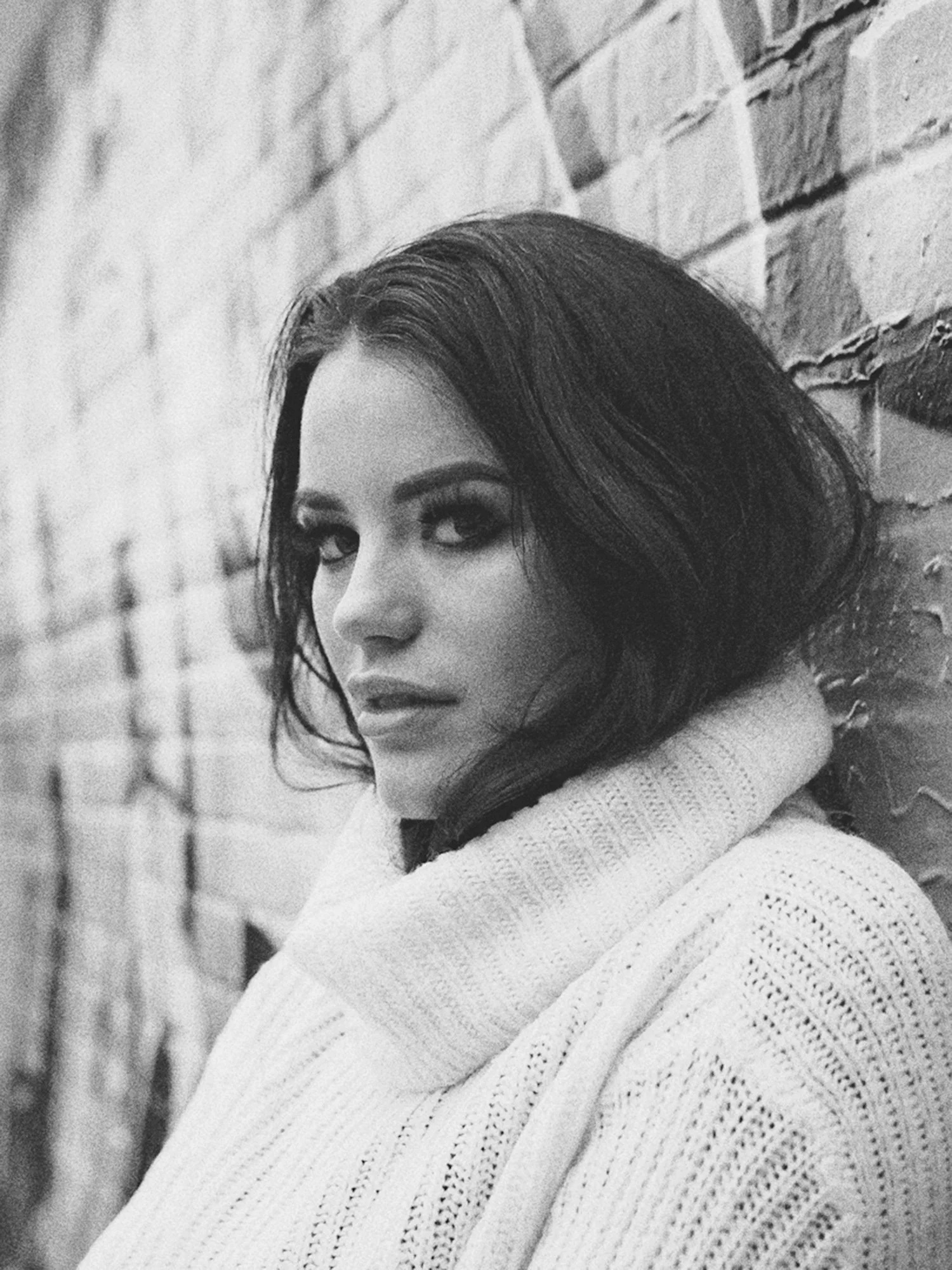 A black and white close-up portrait of a woman with dark, wavy hair, wearing a knitted sweater, leaning against a brick wall and gazing at the camera.