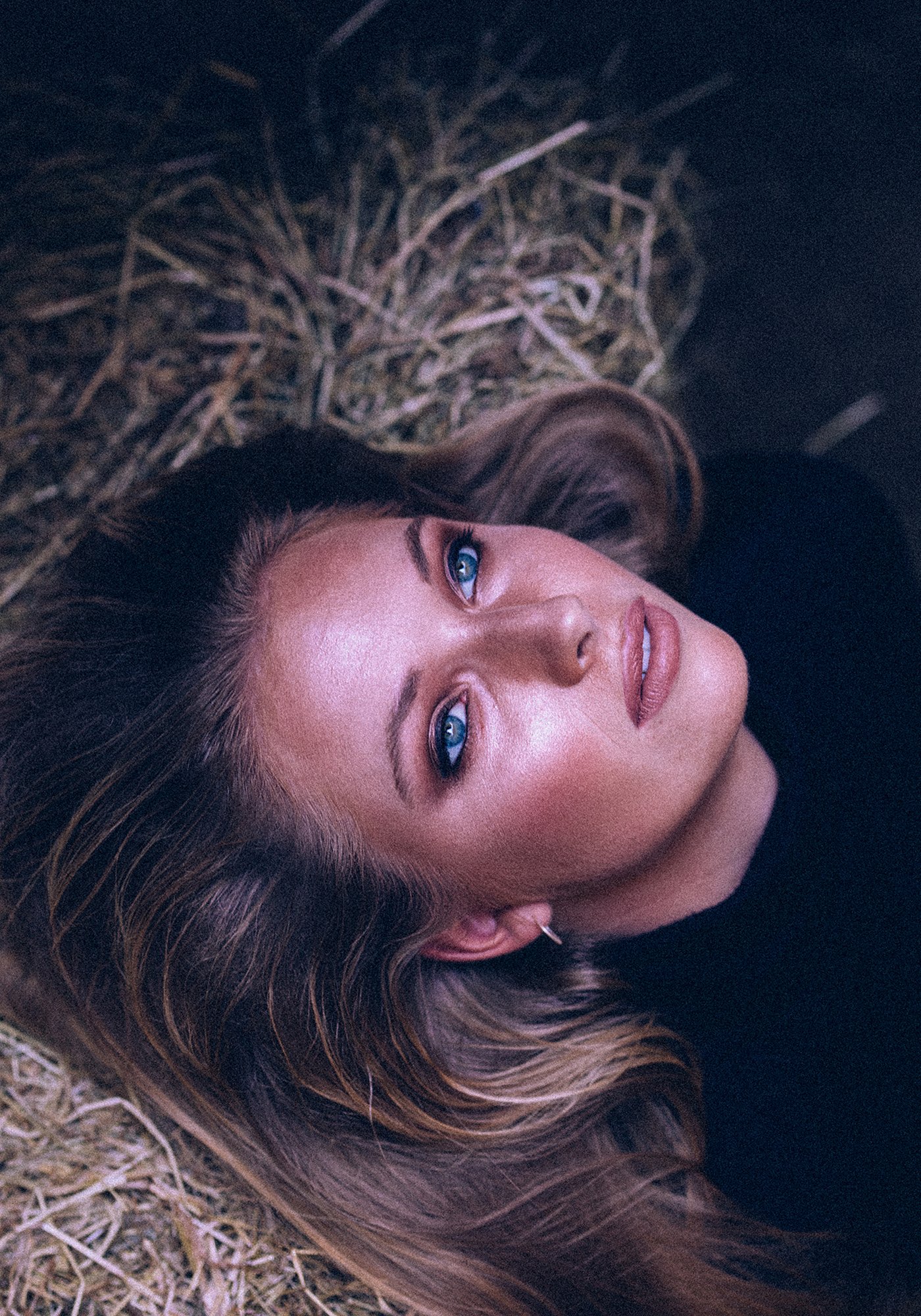 A woman with long, wavy hair and blue eyes lying on a bed of hay, looking up at the camera.