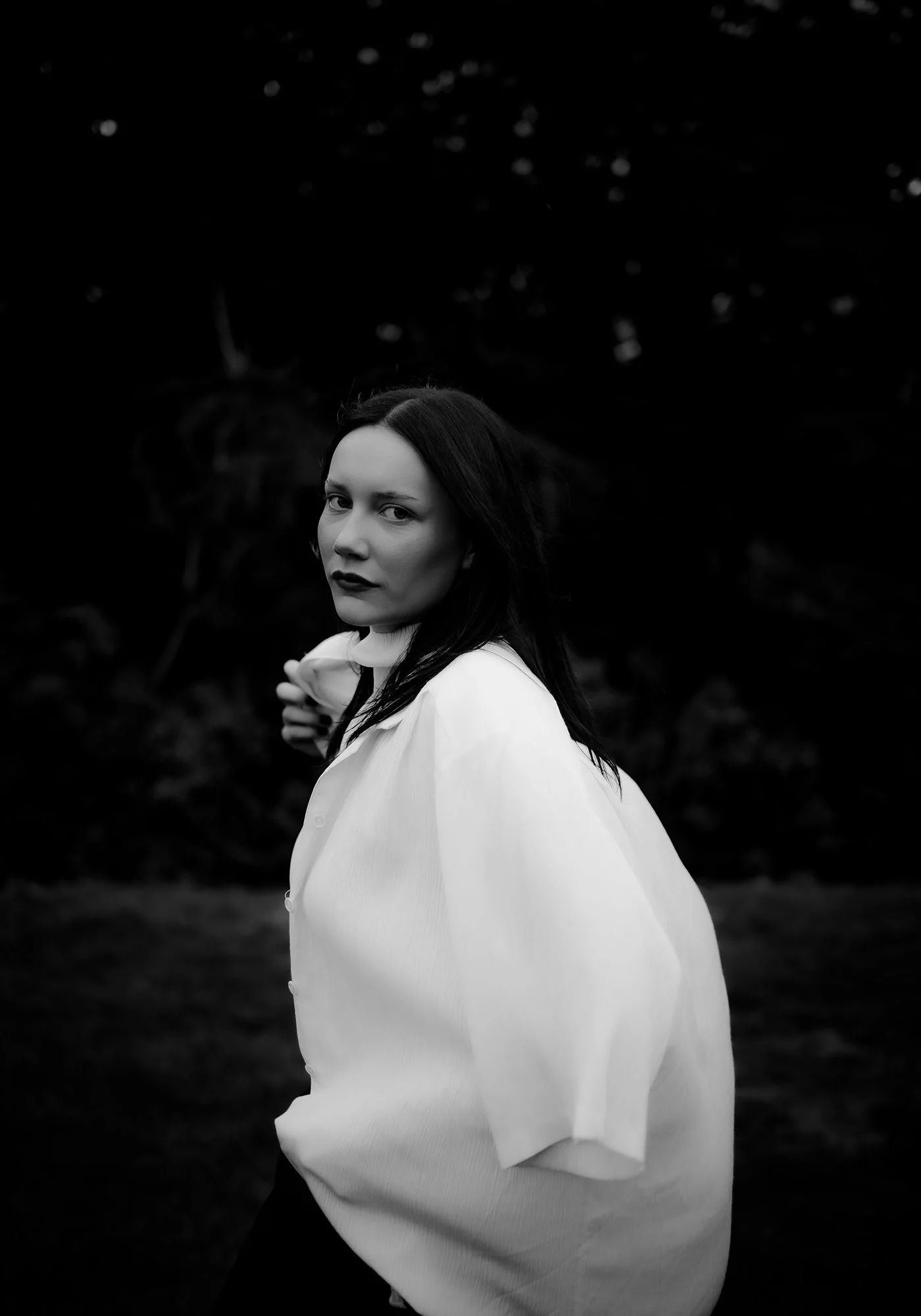 A woman with dark hair and light skin wearing a white blouse standing outdoors in a dark, natural environment, looking at the camera.