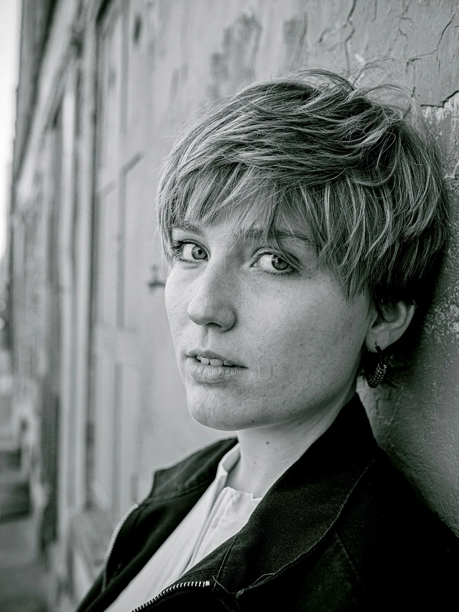 Black and white close-up portrait of a woman with short hair leaning against a brick wall, looking at the camera.