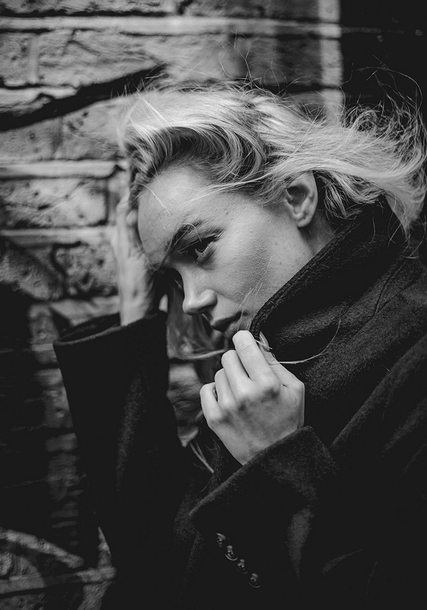 Black and white portrait of a woman with light, wavy hair, wearing a dark coat, posed near a brick wall, partially covering her face with her hand.