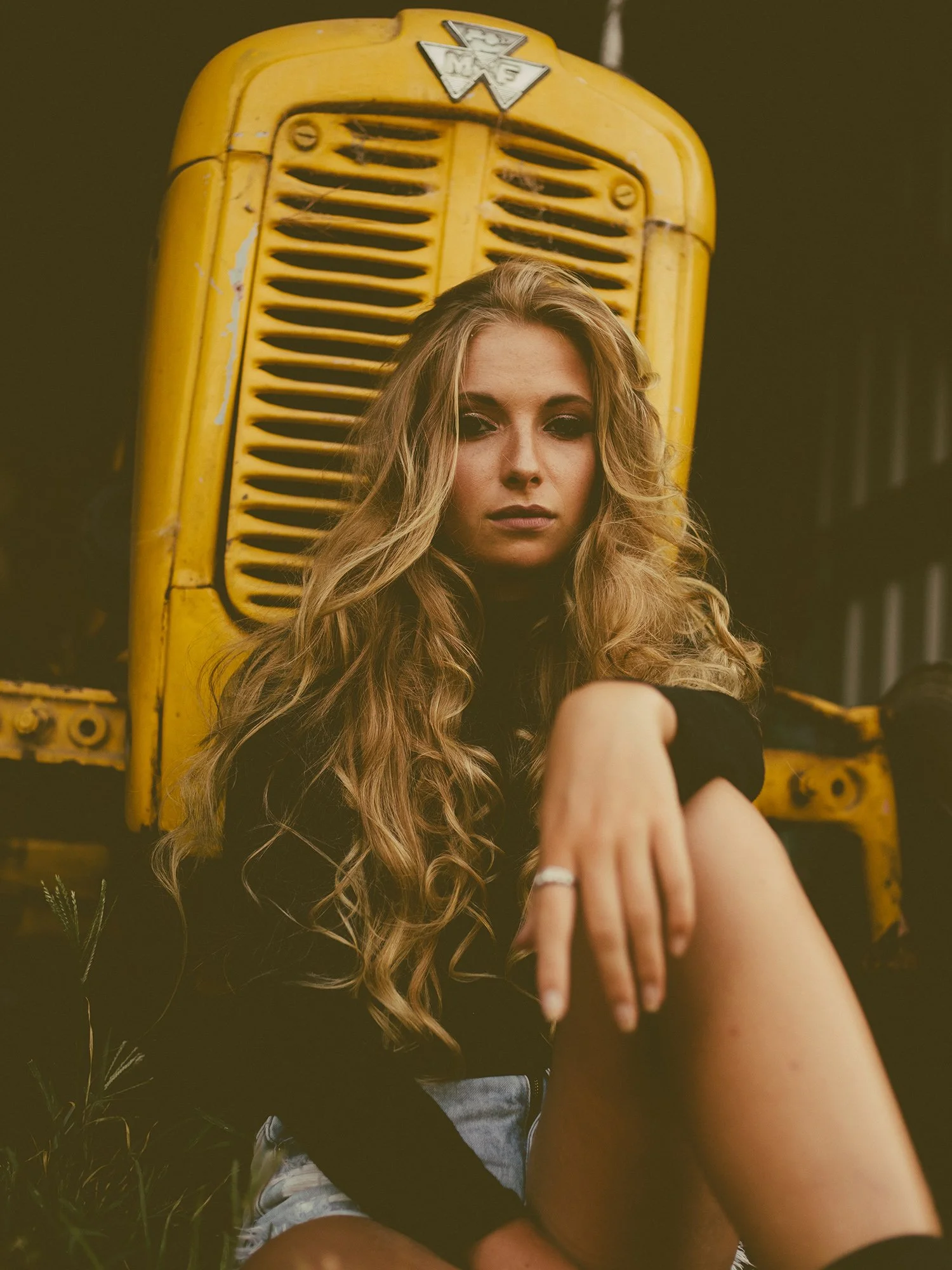 Young woman with curly hair sitting in front of an old yellow vehicle with a grille, wearing a black top and shorts.