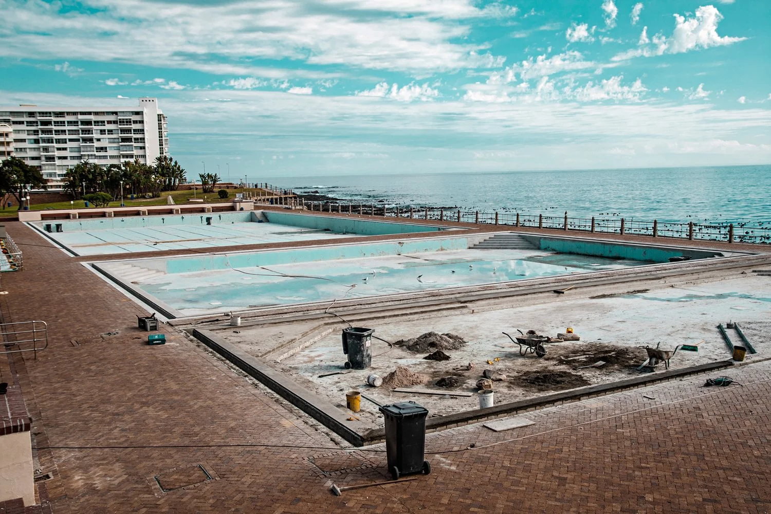 Empty swimming pools under renovation on a beachfront with ocean in the background, construction tools and debris in the foreground, and apartment buildings in the distance.