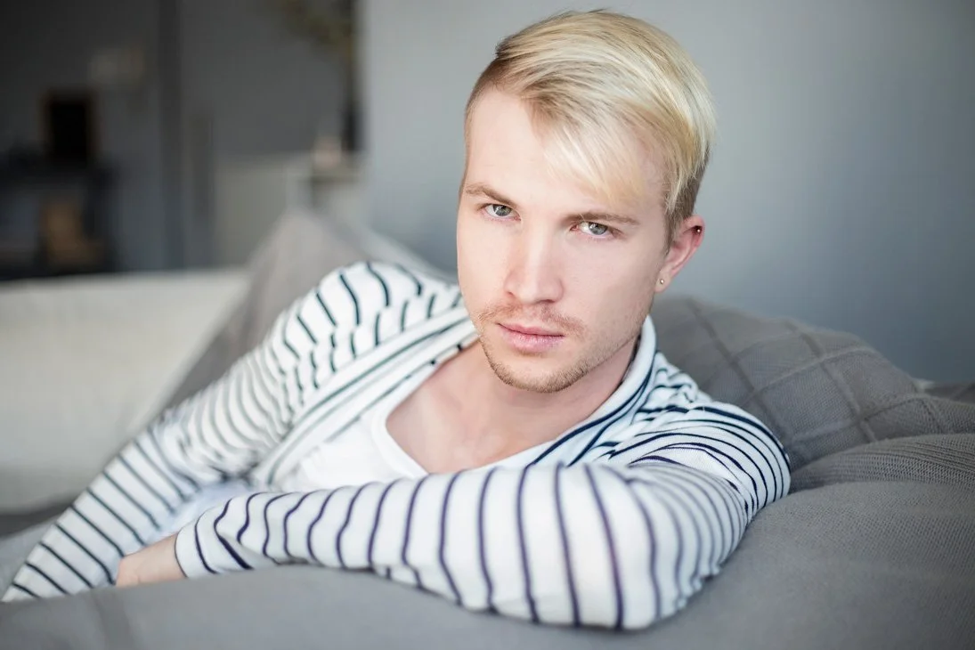A young man with blond hair and light skin laying on a gray couch, wearing a white and black striped shirt, looking directly at the camera.