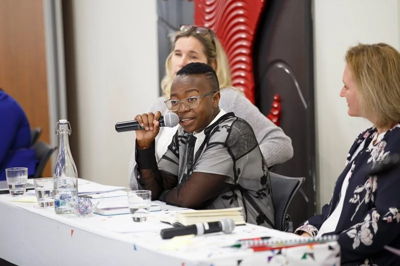 A woman with short black hair and glasses speaking into a microphone at a panel discussion table with other women.