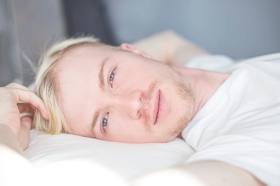 A young man with blond hair and a light beard lying in bed, looking tired or contemplative.