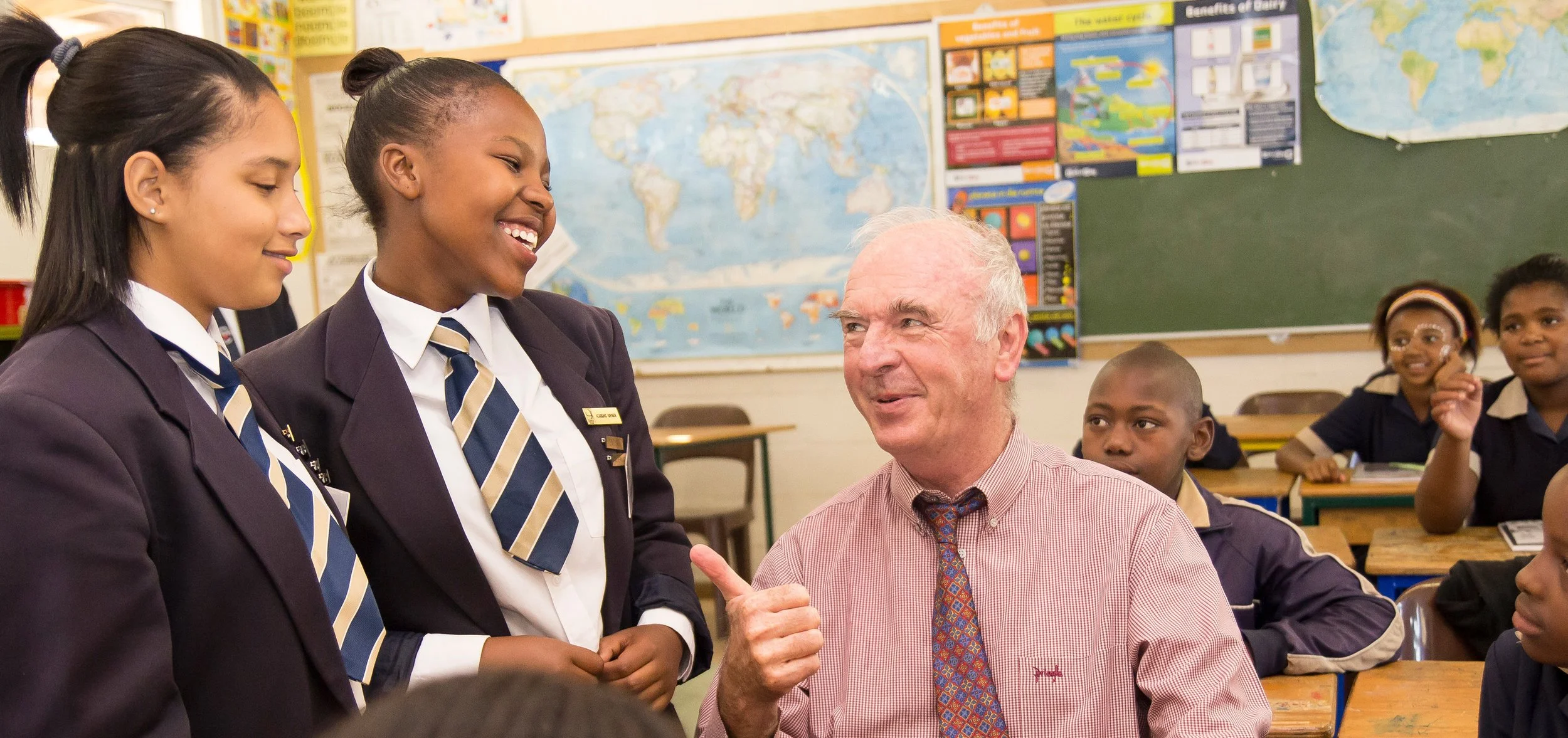 A teacher is engaging with students in a classroom. Two girls are smiling and talking to an elderly man, likely a guest or teacher. Other students are seated at desks in the background, with a world map and educational posters on the classroom walls.
