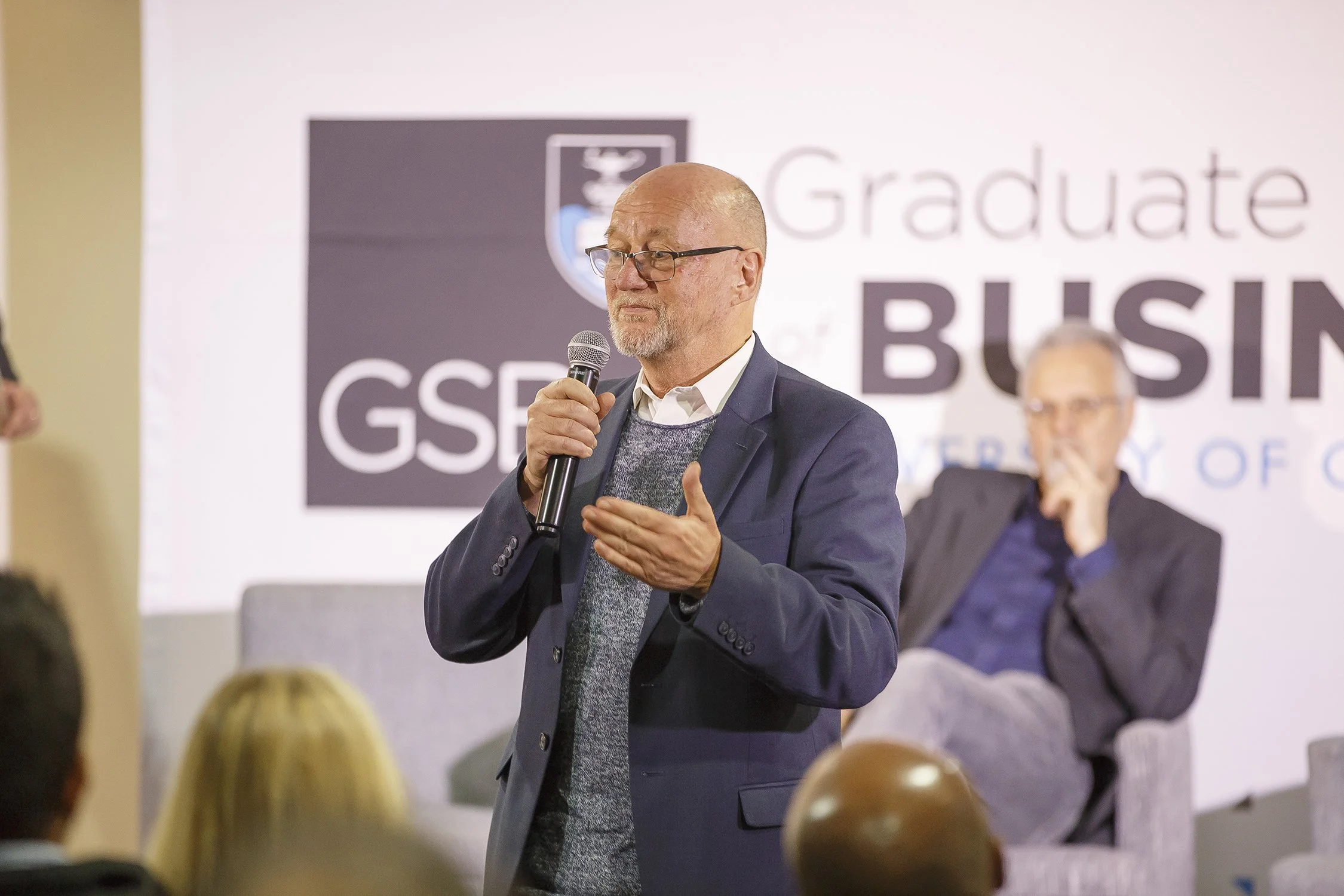 A man with glasses and a beard speaking into a microphone at a GSB graduate business event, with a blurred seated man in the background and an audience in front.