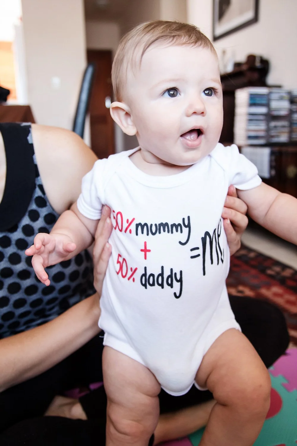 A baby wearing a white onesie with fun math-related text and symbols, held up by an adult.
