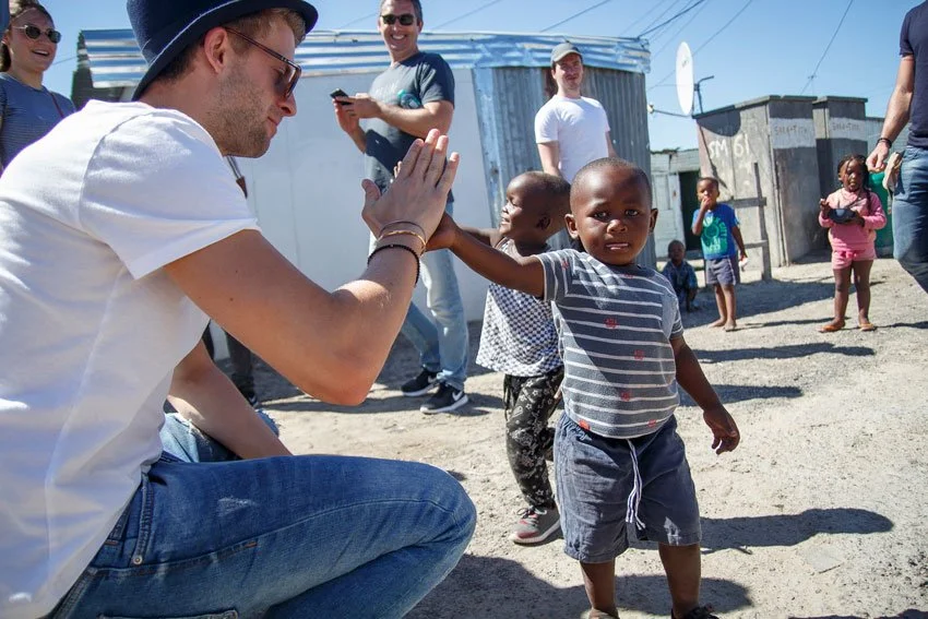 A man squatting down giving a high five to a young boy in a striped shirt in a rural setting. Others, including children and adults, are standing or playing nearby, with simple metal houses and a satellite dish in the background.