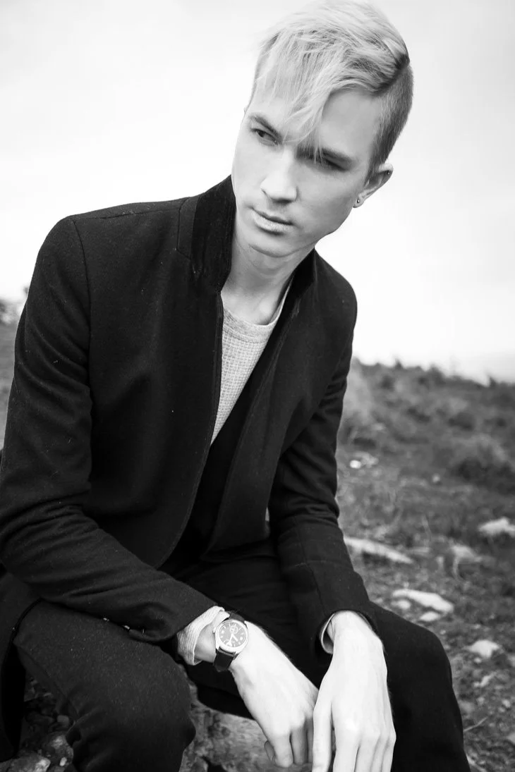 Black and white photo of a young man with styled blonde hair, sitting outdoors on a rock, wearing a black jacket, a watch, and casual clothing, looking off to the side.