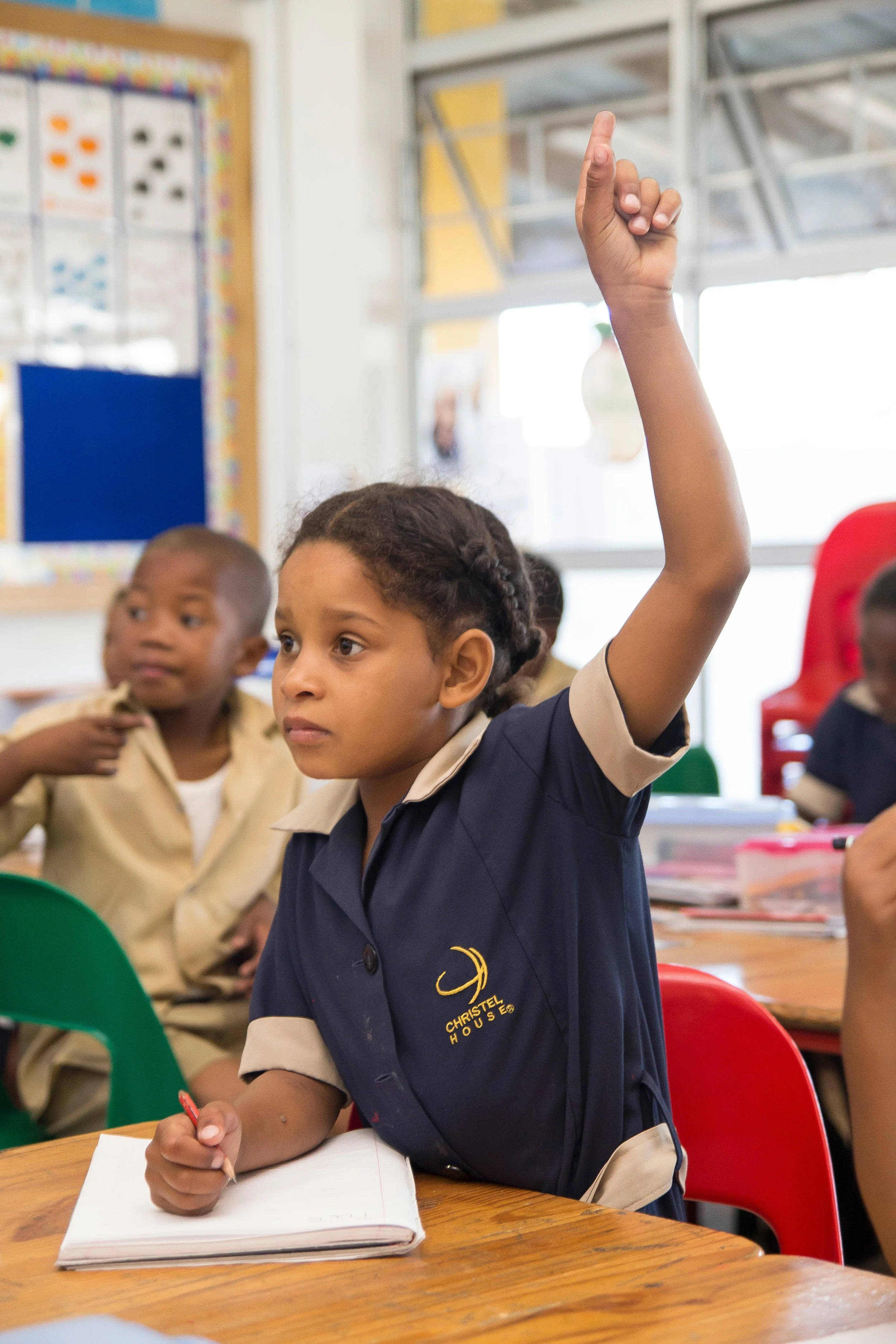 A young girl in a school uniform raising her hand in a classroom, with other children seated around her.
