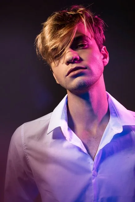 Portrait of a young man with blonde hair, wearing a white shirt, illuminated by pink and purple lighting.