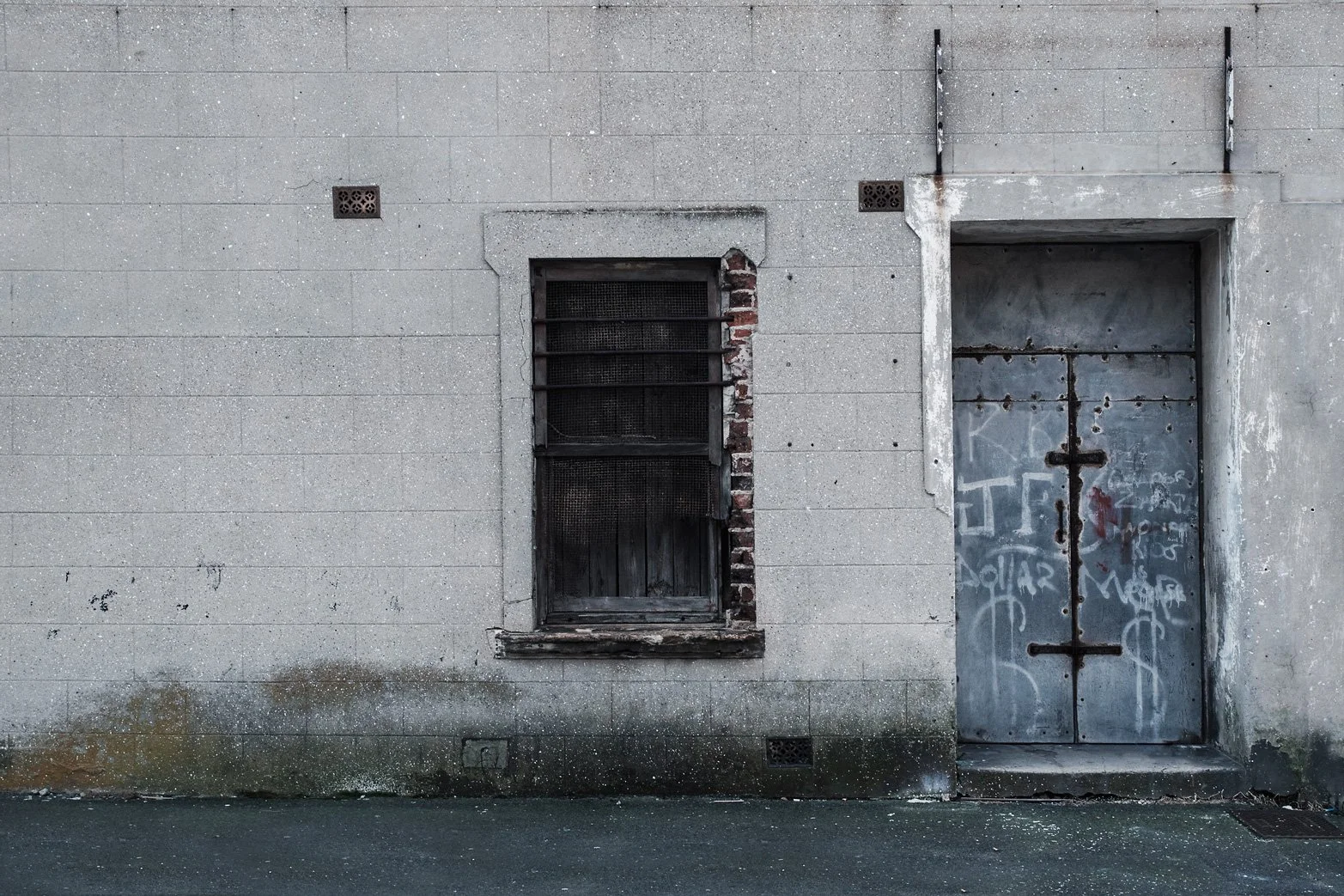 A weathered concrete building wall featuring a barred window with brick and wood framing, and a rusted metal door with graffiti, set above a damp, stained sidewalk.