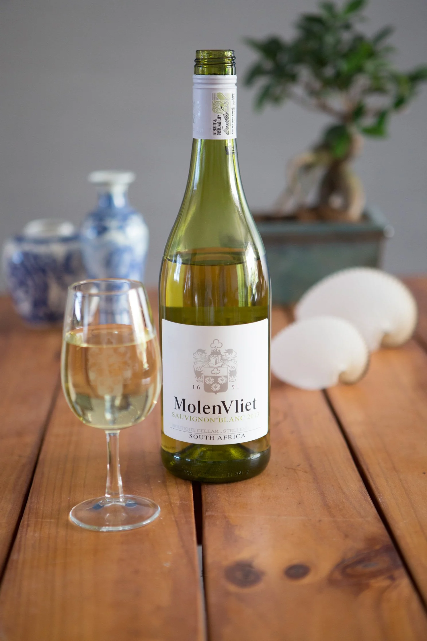 A bottle of MolenVliet Sauvignon Blanc white wine with a glass of poured wine on a wooden table, with decorativeware and a bonsai plant in the background.
