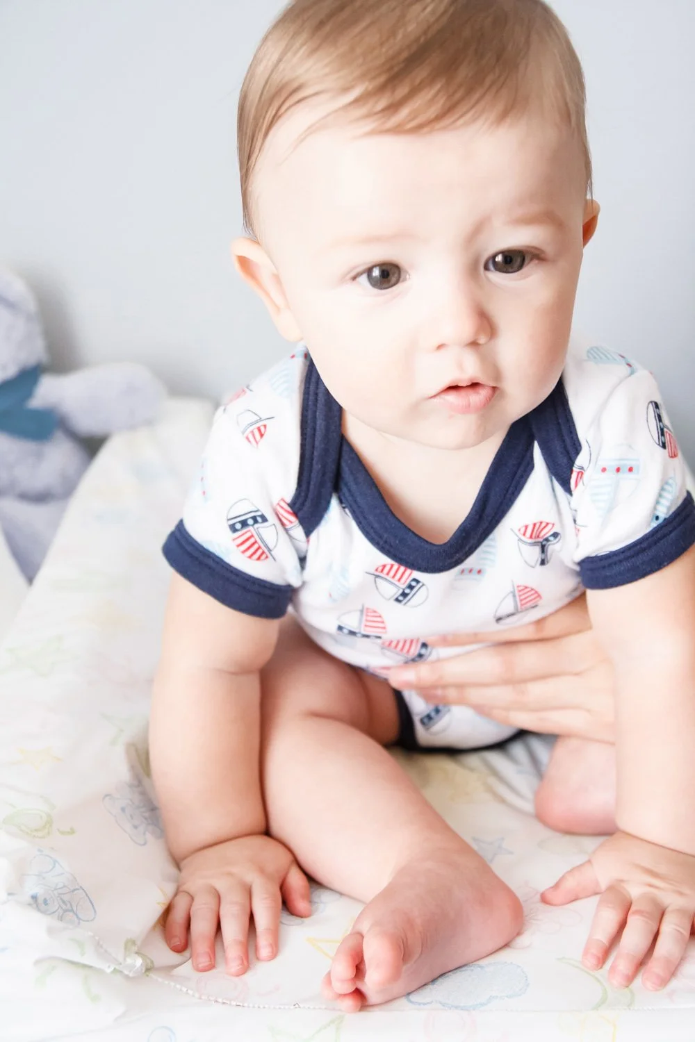 A young baby with light brown hair and light skin, wearing a white shirt with navy and red patterns, sitting on a bed or changing table.