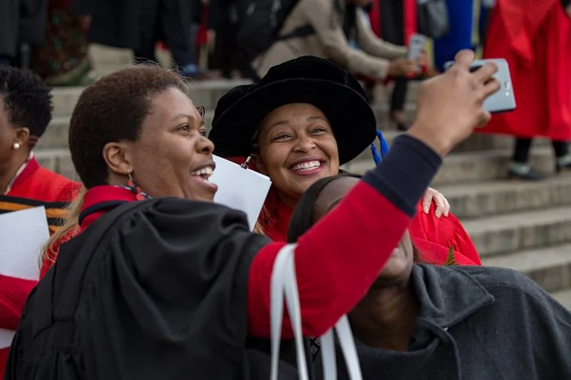 Two women with short hair and a third person with a dark hat are taking a selfie together on a set of steps, with other people in the background.
