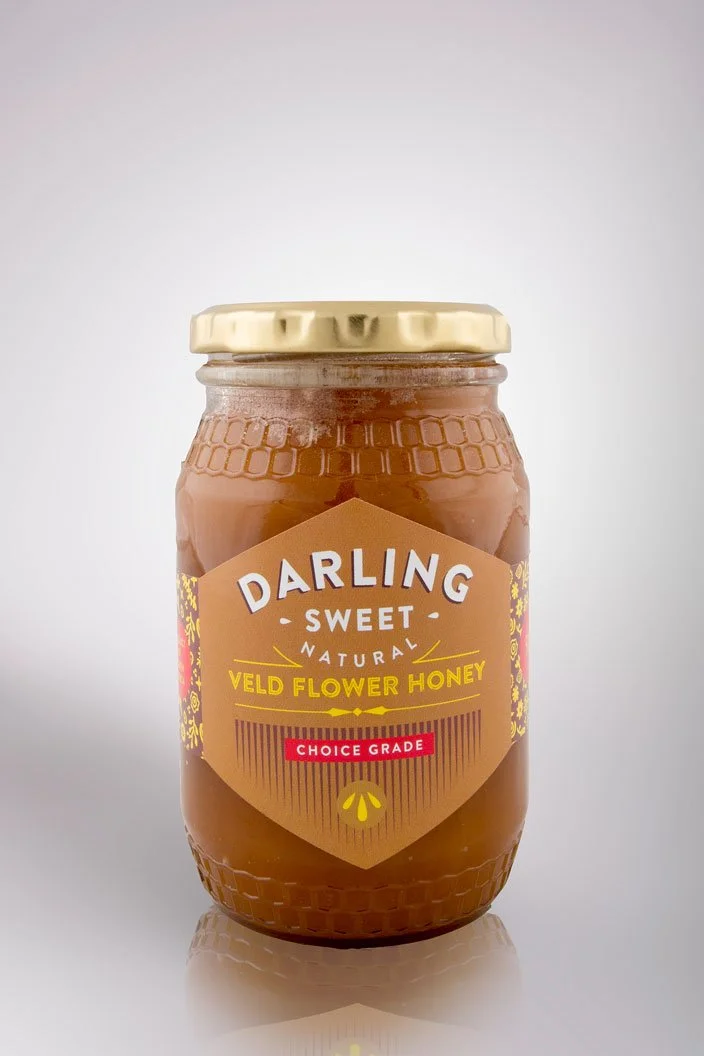 A jar of Darling Sweet natural veld flower honey with a beige lid, placed on a reflective surface against a plain background.