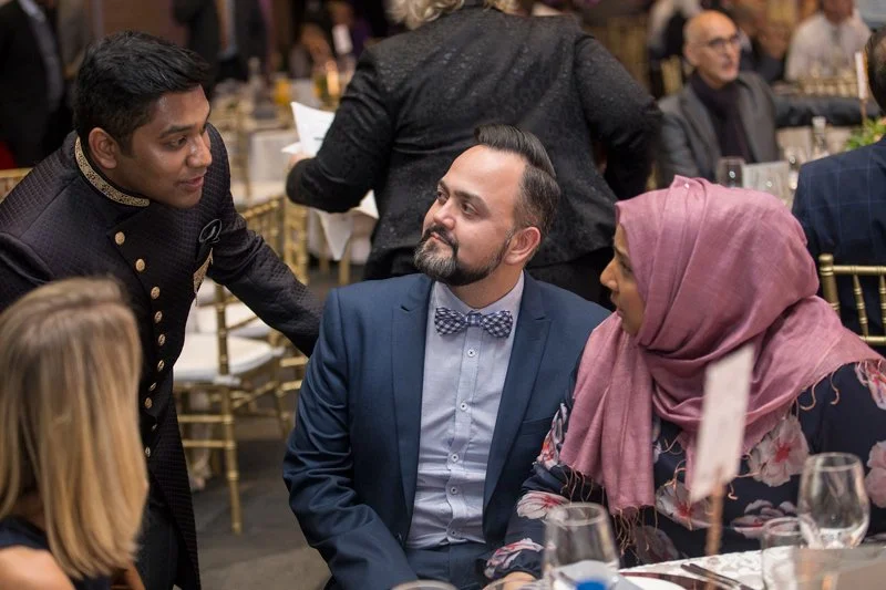 Man in a blue suit with a bow tie engaged in conversation at a formal event, with a woman in a pink hijab sitting nearby and another man in traditional attire standing and talking to him.