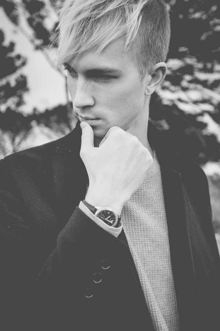 Black and white photo of a young man with blonde hair, wearing a watch, a textured shirt, and a dark blazer, looking thoughtful with his hand on his chin.