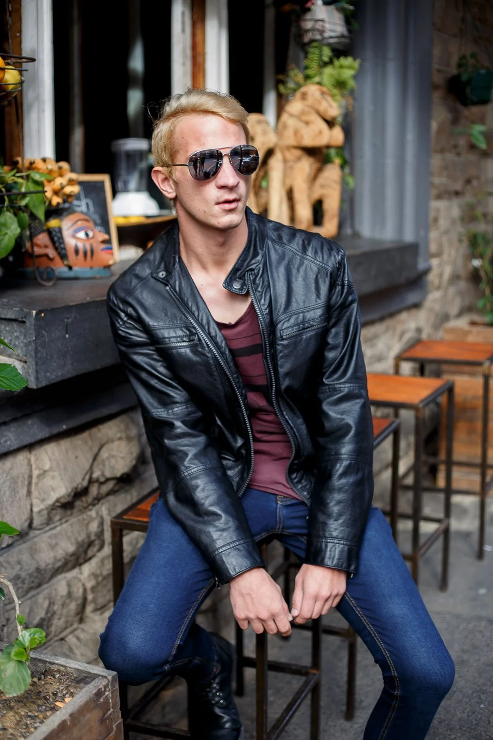 A young man with blonde hair wearing sunglasses, a black leather jacket, a maroon shirt, and blue jeans, sitting outdoors on a metal stool near a stone wall with plants and decorative items.