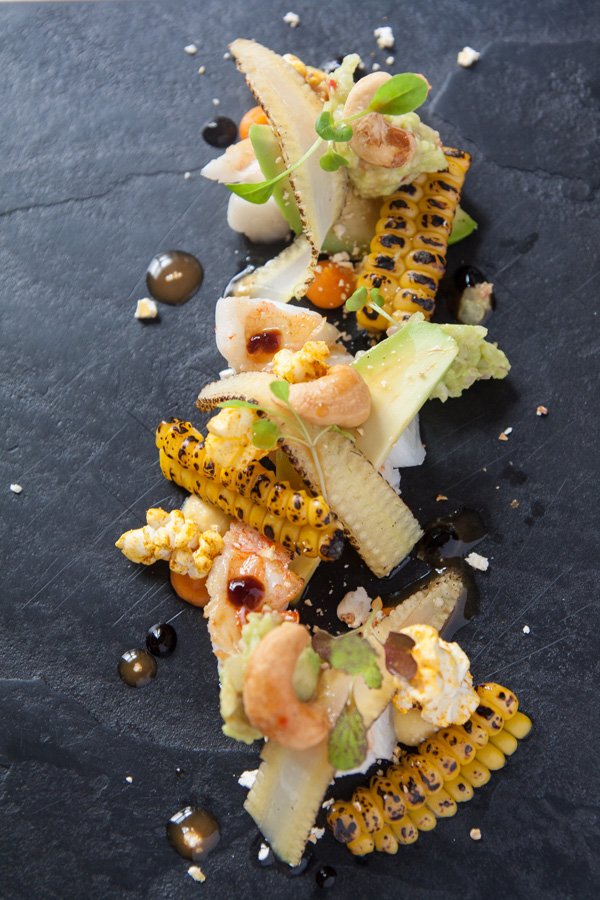 Gourmet dish with grilled corn, slices of fish, microgreens, and drizzled sauces on a black slate plate.