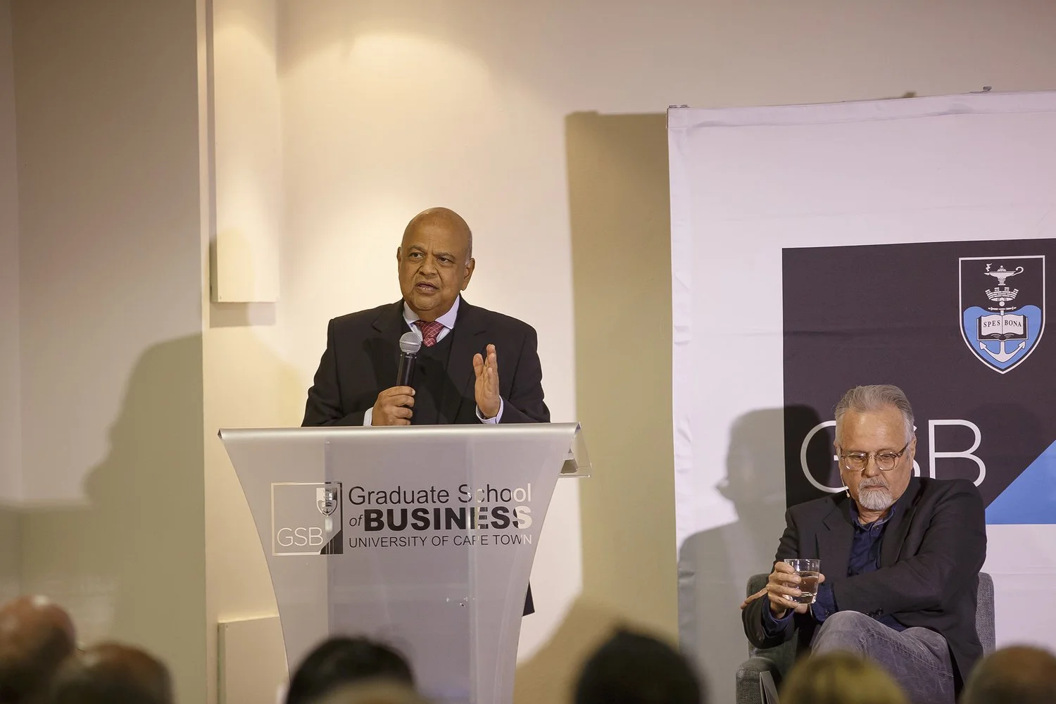 A man in a suit speaking into a microphone at a podium labeled Graduate School of Business, University of Cape Town, with a man seated to his right holding a glass of water. Audience members are visible in the foreground.