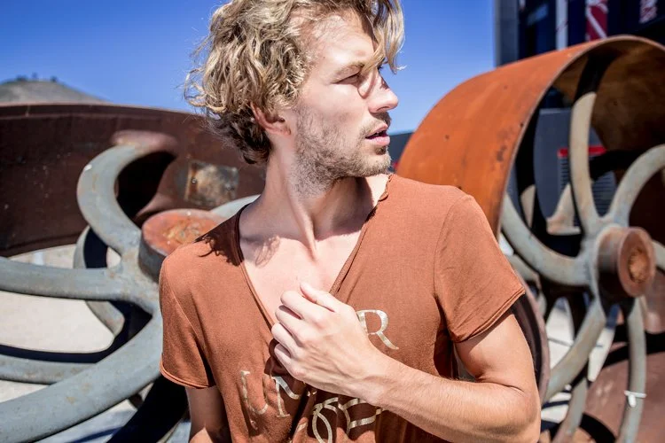A young man with curly blonde hair and a light beard, wearing a brown shirt, stands outdoors surrounded by old, rusted metal equipment, possibly farming or industrial machinery, under a blue sky.