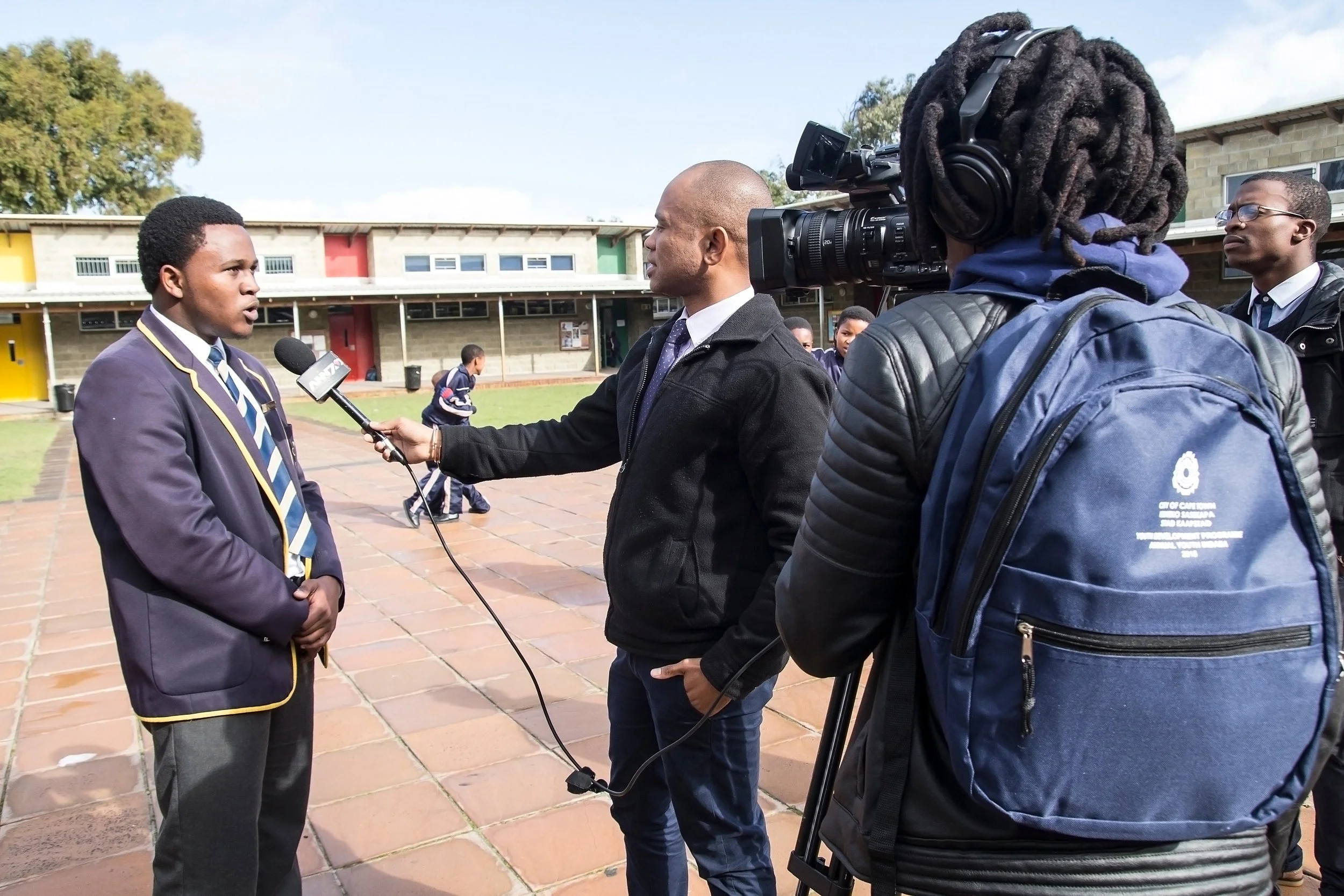 A young male student in a school uniform is being interviewed by a news reporter holding a microphone at a school yard, with another person recording the interview with a camera.