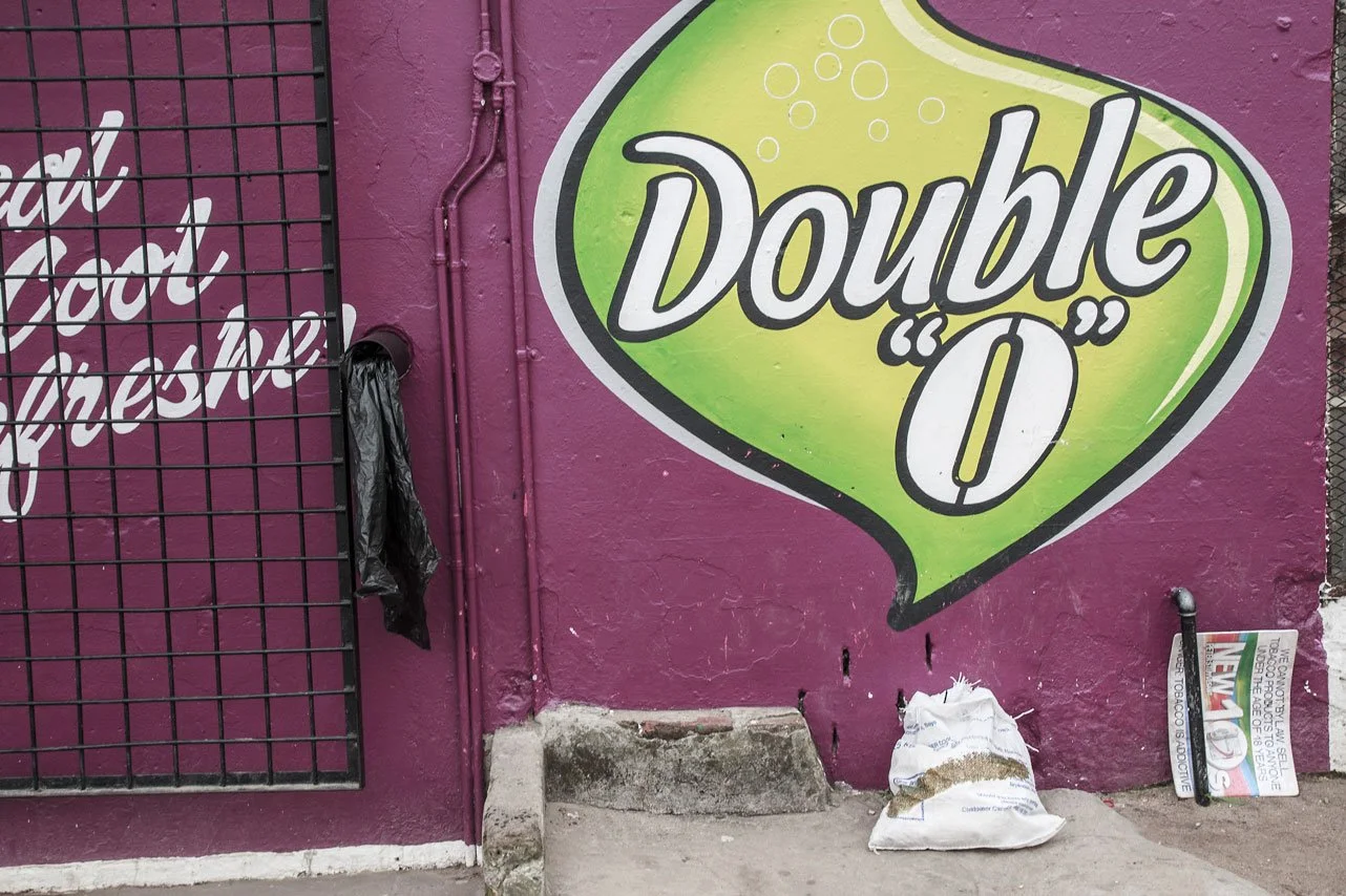 A mural on a purple wall advertising 'Double O' with a green lemon-lime design behind the text. A black metal gate is on the left, with a black trash bag hanging from it. There's a small pile of a white sack and a partially visible political sign on 
