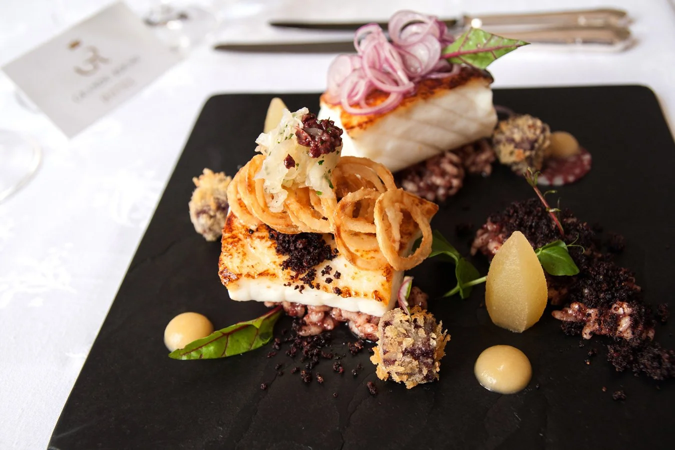 Gourmet dish with seared fish, crispy onion rings, pickled red onions, microgreens, and various sauces on a black slate plate.