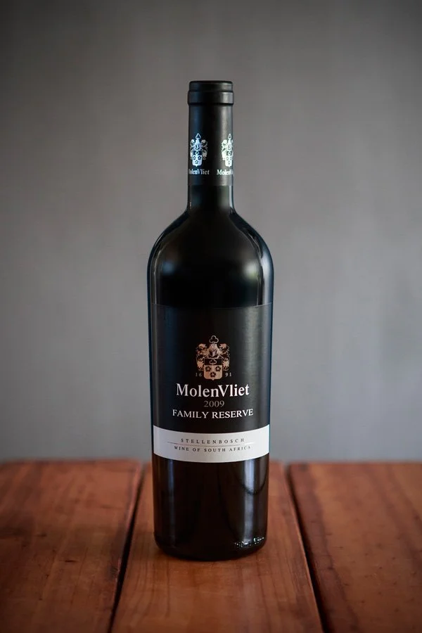 A dark bottle of MolenVliet Family Reserve wine from Stellenbosch, South Africa, placed on a wooden surface with a plain gray background.