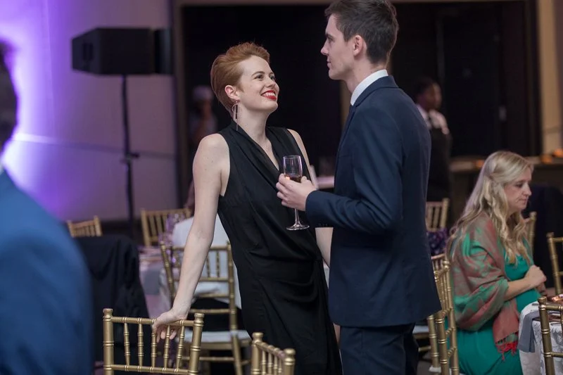 A woman in a black dress and a man in a suit are smiling and looking at each other while talking at an event. The woman is holding a glass of wine.