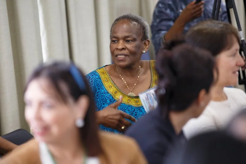 An older woman with short gray hair wearing a blue and yellow dress is speaking to a group of women in a conference or meeting setting.