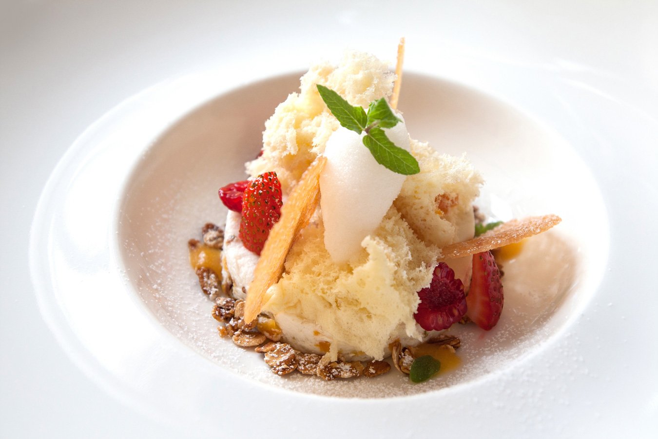 Decorative dessert with strawberries, mint leaf, vanilla ice cream, granola, and edible decorations on a white plate.